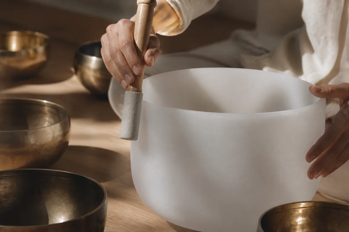 Hands circling a mallet around the rim of a frosted crystal singing bowl, brass bowls nearby